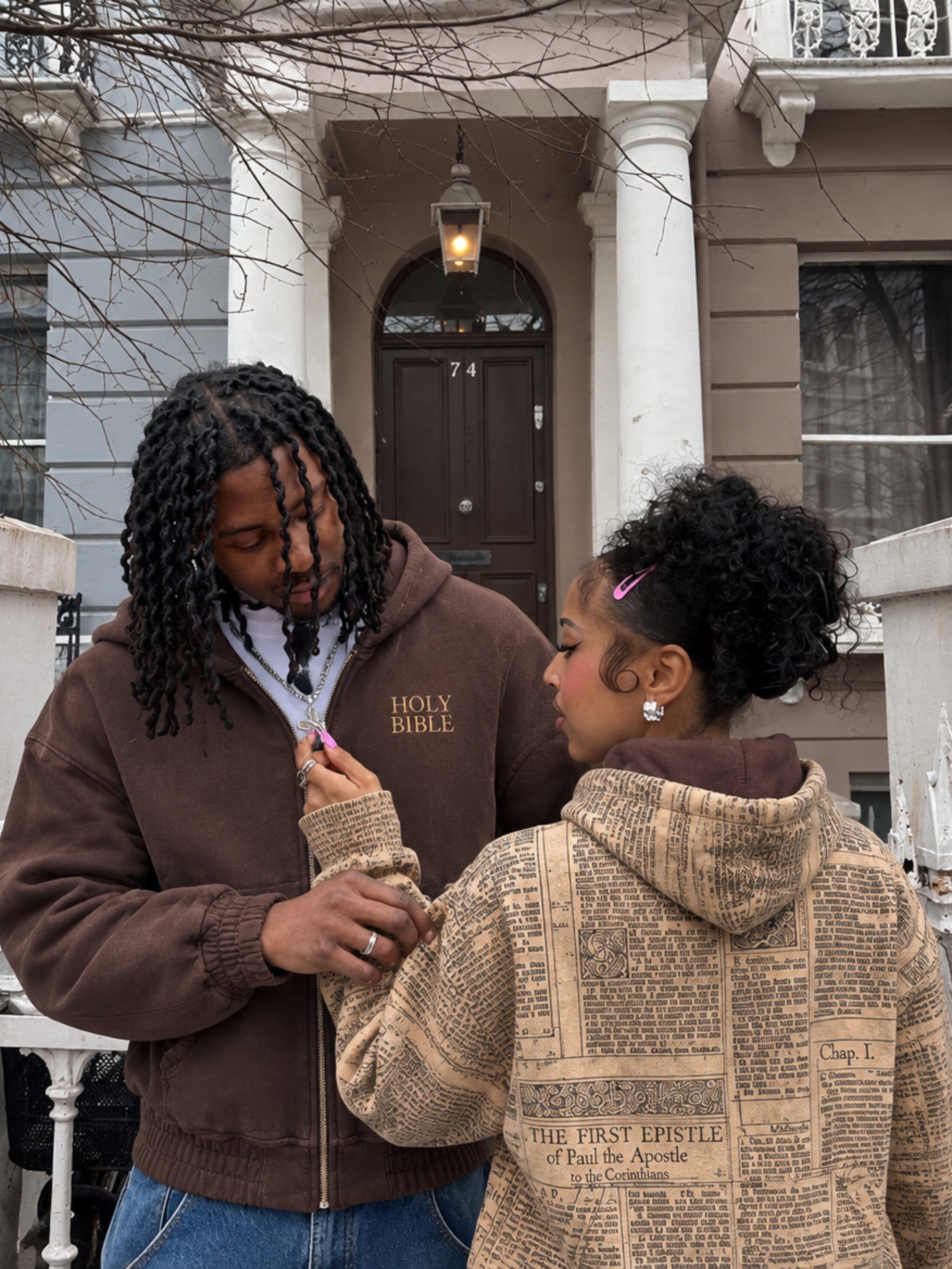 A couple outside a townhouse wearing both sides of the HOLY Reversible Zip-Up: chocolate brown with HOLY BIBLE print, and the vintage scripture print