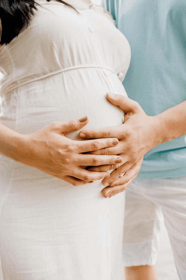 Schwangere Frau in weißem Kleid, die mit beiden Händen den Bauch hält, die Hände des Partners in blauem Hemd darüber.