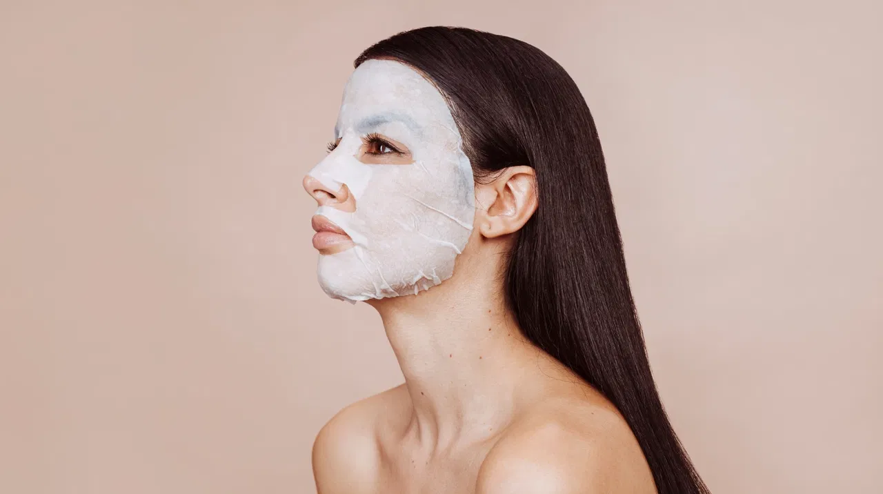 Woman with long hair wearing a sheet mask, facing right on a beige background. Verjüngt Kollagen die Haut?