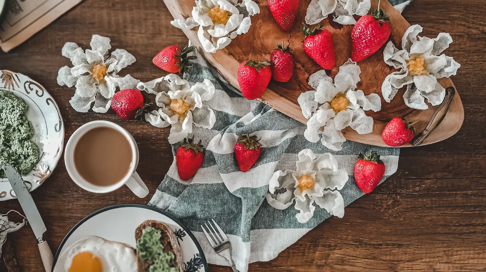 Rustikaler Frühstückstisch mit frischen Erdbeeren, Eiern, Kaffee und grünen Aufstrichen, dekoriert mit Blumen.