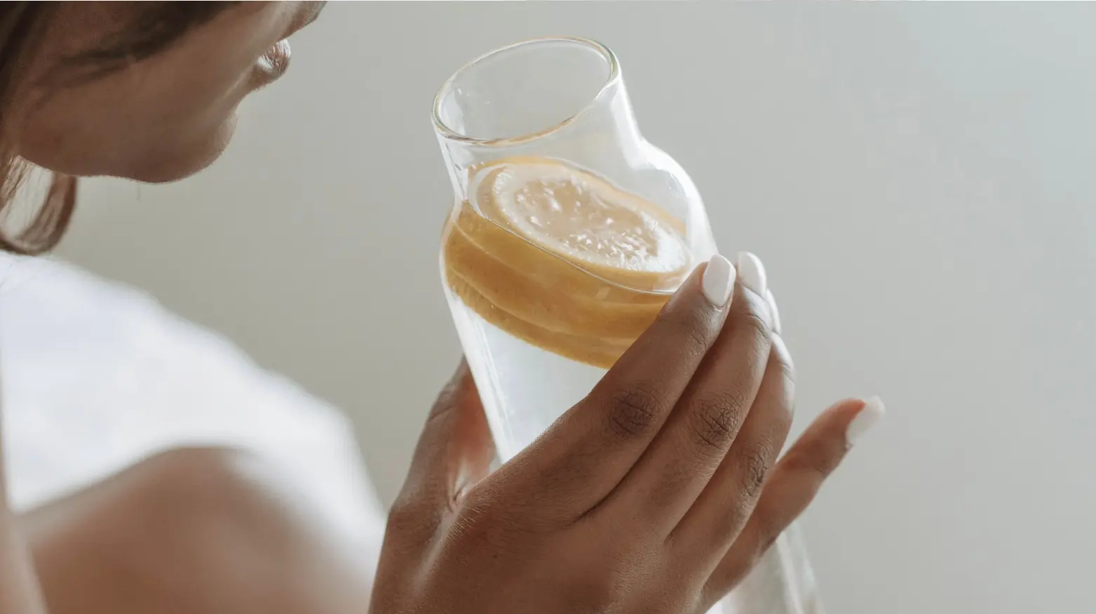 Nahaufnahme einer Person, die ein Glas Wasser mit Zitronenscheiben hält, Hand mit weiß lackierten Nägeln im Fokus.