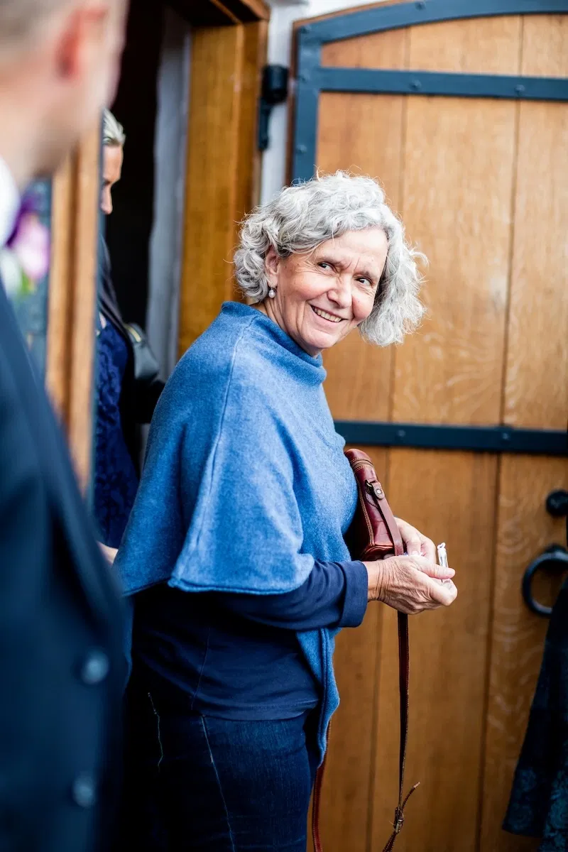 Eine lächelnde Frau mit grauen Haaren und blauem Schal steht an einer Holztür, in der Hand eine Handtasche und Schlüssel.