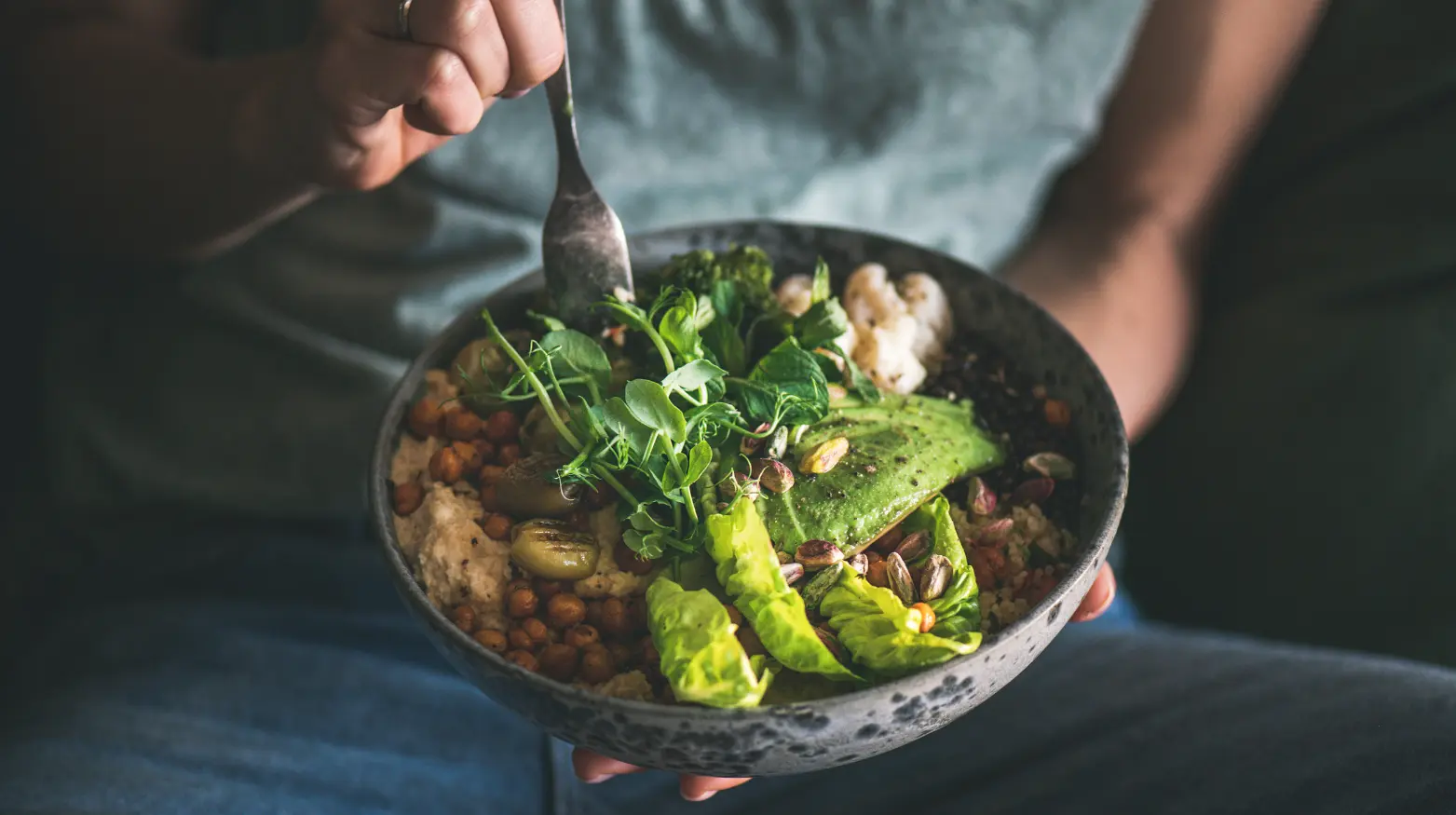 Eine Schale mit Avocado, Grünzeug, Kichererbsen, Pistazien und Hummus, die von einer Person mit einer Gabel gehalten wird.