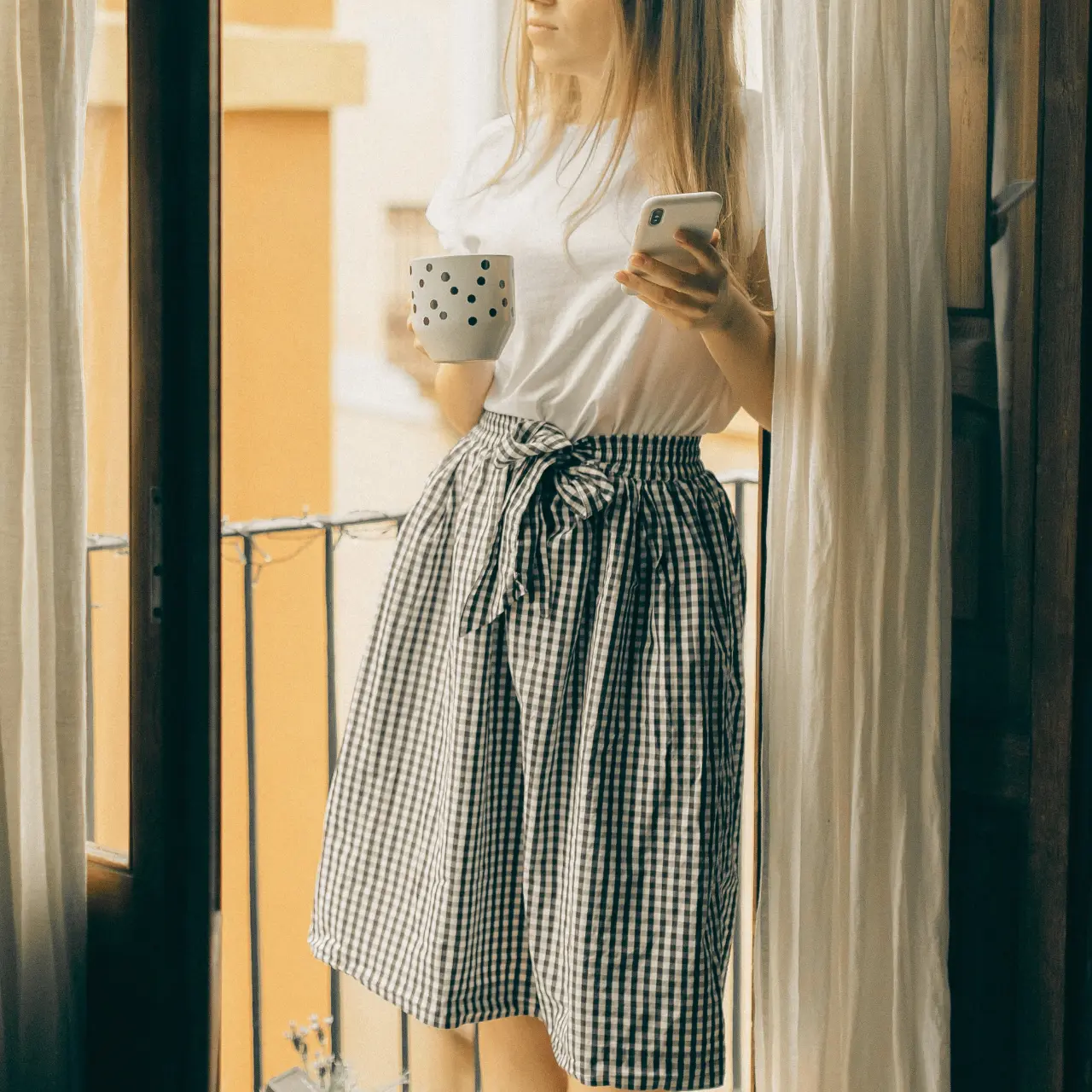 Eine Frau in einem karierten Rock hält einen gepunkteten Becher und ein Smartphone in der Hand und steht an einem Fenster mit Vorhängen.