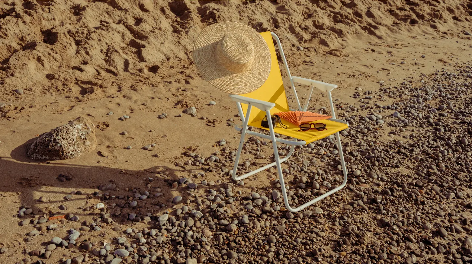 Ein gelber Strandstuhl mit Strohhut und Sonnenbrille steht auf einem Kieselstrand.