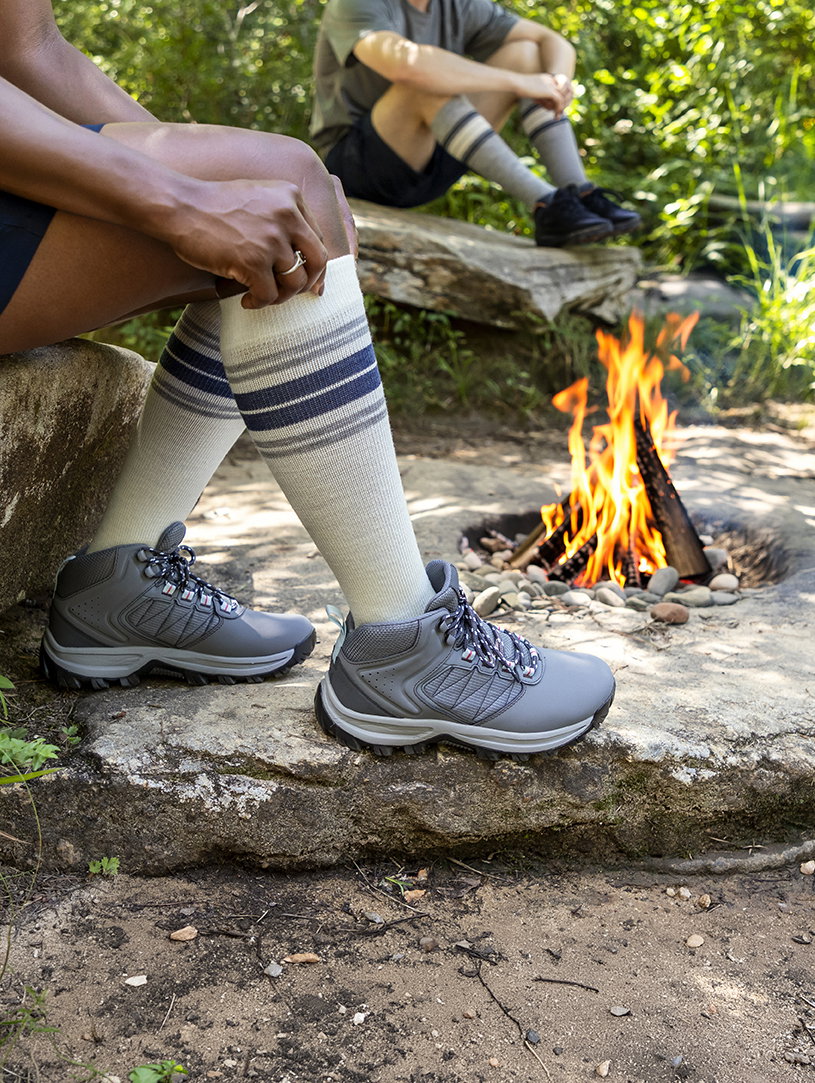Two campers resting on rocks beside a campfire in the woods, wearing Cream and Heather Grey Multi-Stripe Motion Thermoregulating Wool compression socks with hiking boots
