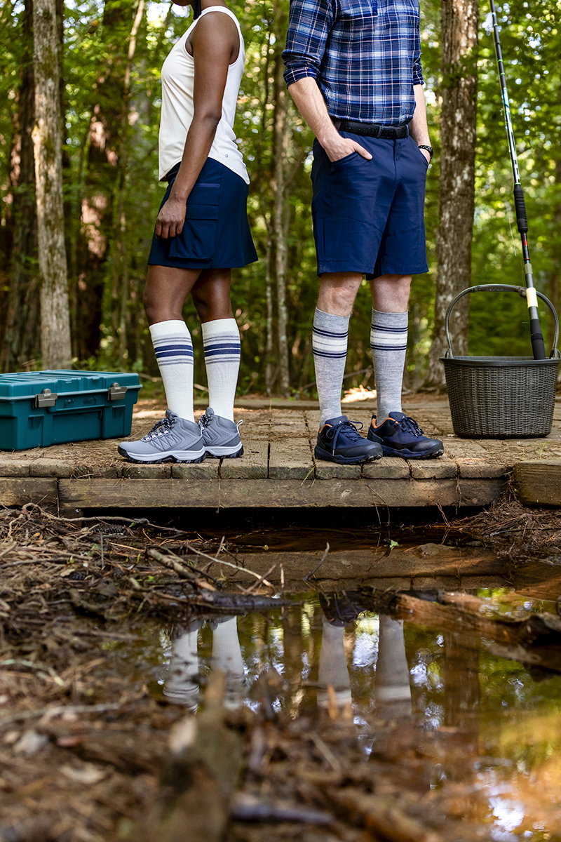 Couple standing on a wooden footbridge over a forest creek with a fishing rod, tackle box and basket, wearing Cream and Heather Grey Multi-Stripe Motion Thermoregulating Wool compression socks