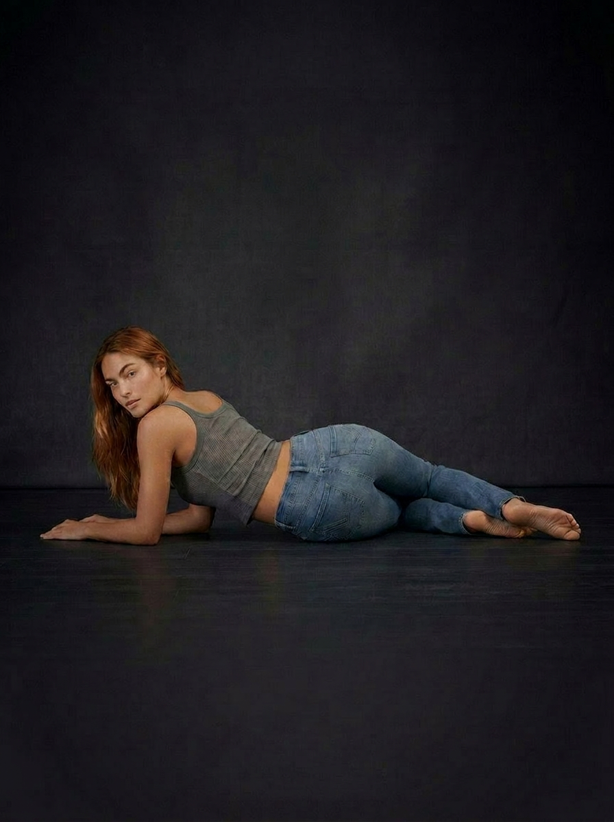 A woman with red hair lying on her side on a dark floor, wearing a grey tank top and blue jeans.