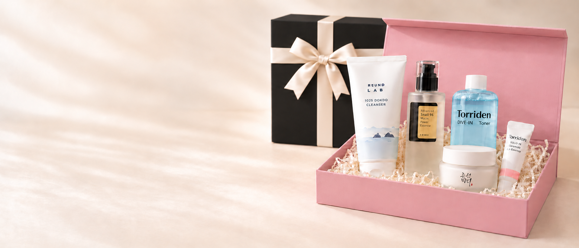 Open pink gift box filled with Korean skincare from Round Lab, COSRX, Torriden and Beauty of Joseon, beside a black gift box tied with a cream satin ribbon