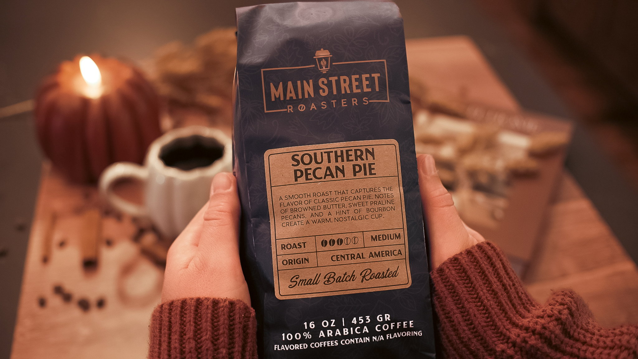 Hands holding a bag of Southern Pecan Pie coffee beside a candle and a warm mug of coffee