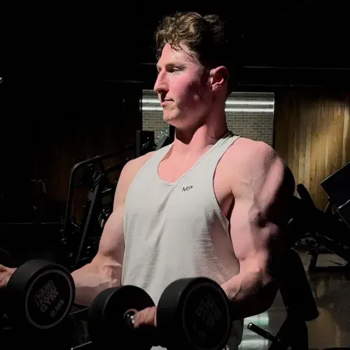 Man in a white tank top lifting dumbbells in a dimly lit gym.