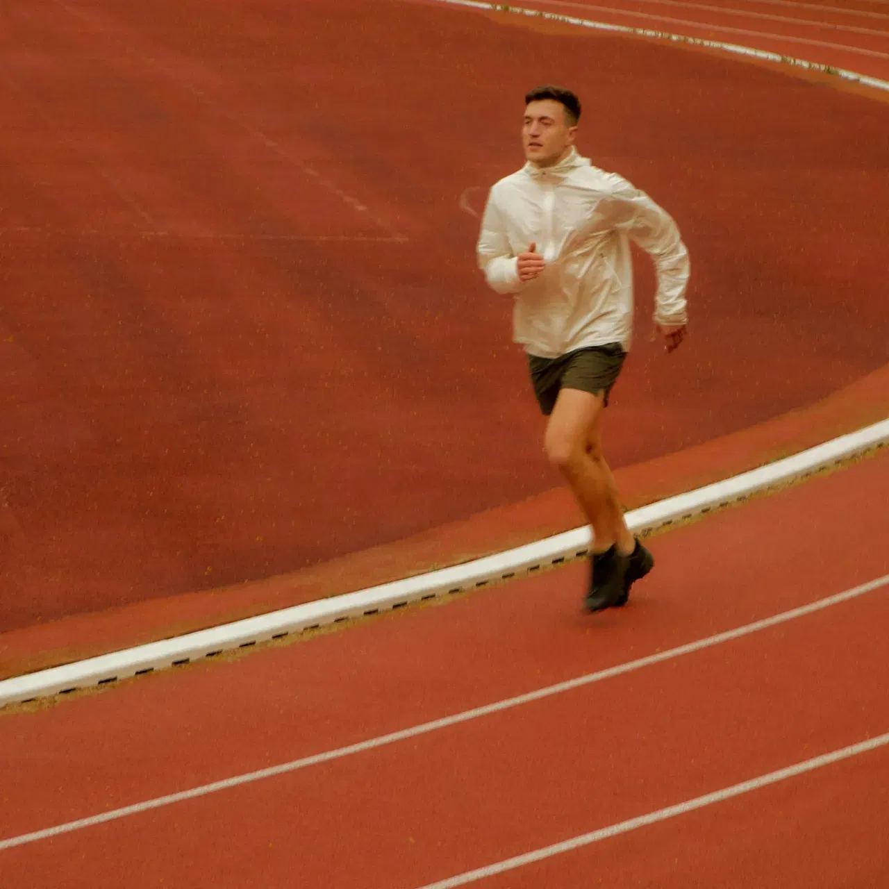 Man running on a curved outdoor red track in a white jacket and black shorts.