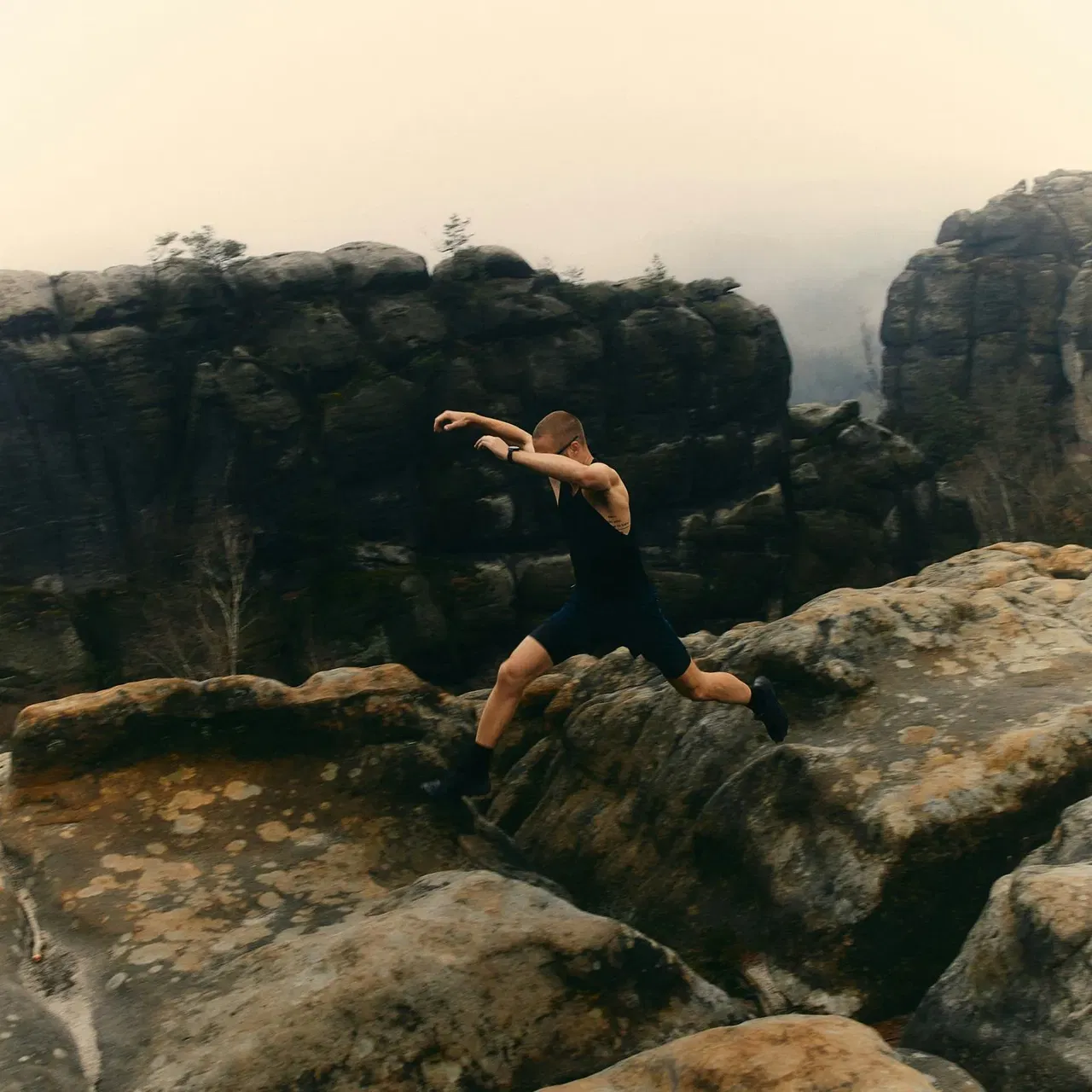 Person in black athletic wear jumping between rocky cliffs with large rock formations in background.