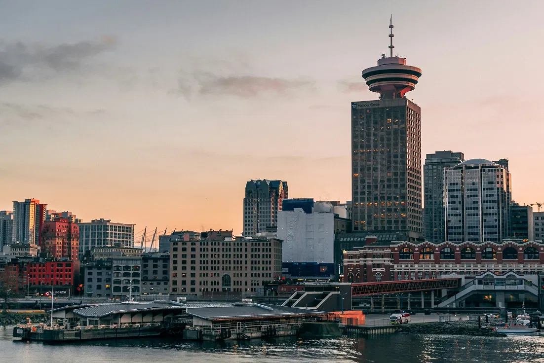 City skyline at dusk with a prominent tower and waterfront buildings under a pastel sky.