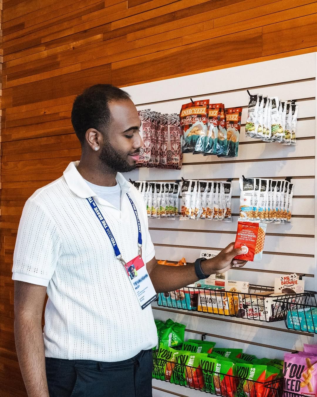 Man in white shirt holds a maple terroir strawberry stroopwafel, standing by a wall of assorted packaged snacks.