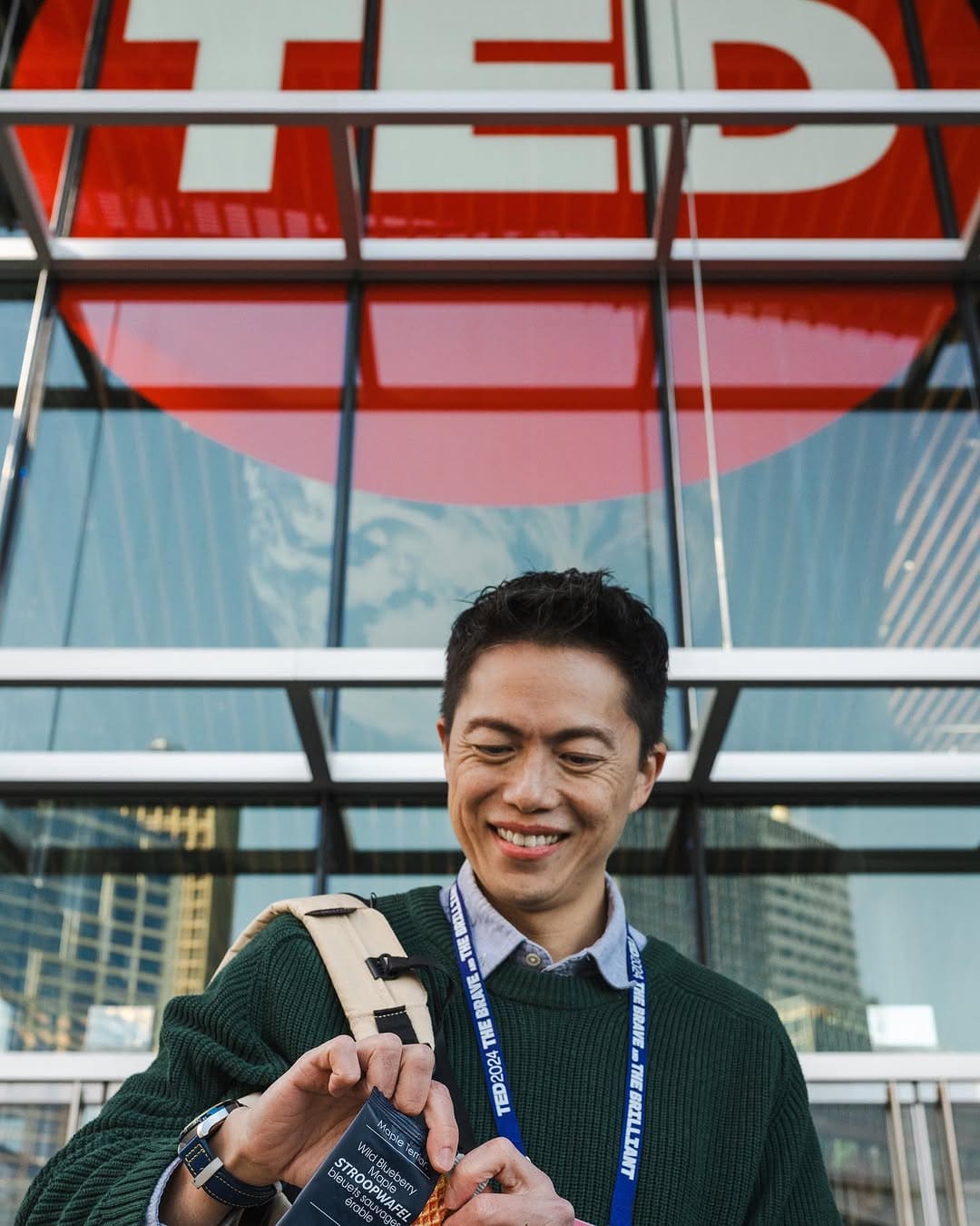 
                        Person smiling with a backpack in front of a glass building with a large TED logo holding a blueberry Maple T
                        erroir stroopwafel.
                      