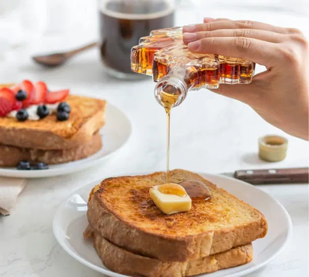 Syrup poured onto buttered French toast with berries in the background.