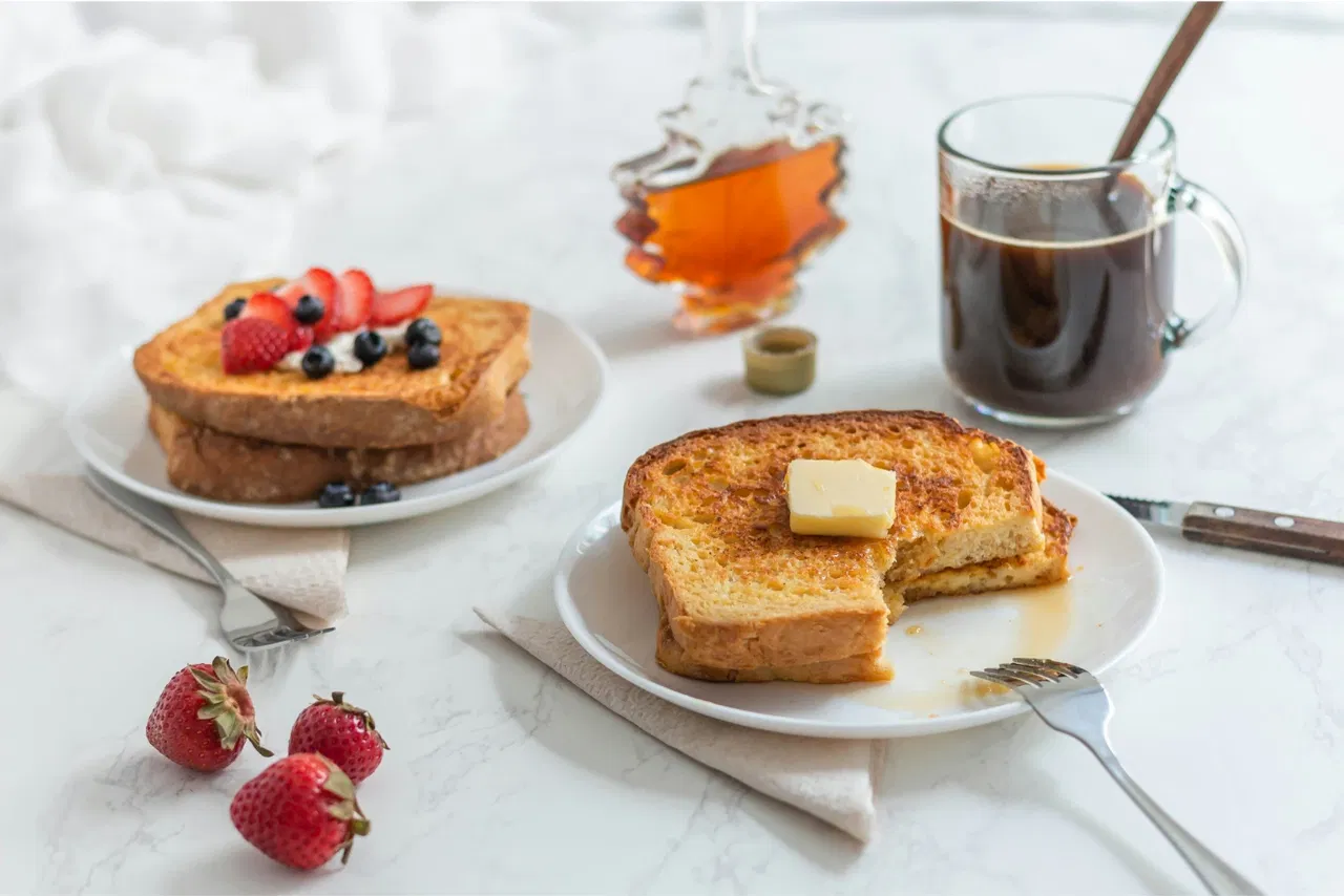 French toast with butter and berries, syrup bottle, coffee cup, and strawberries on a table.