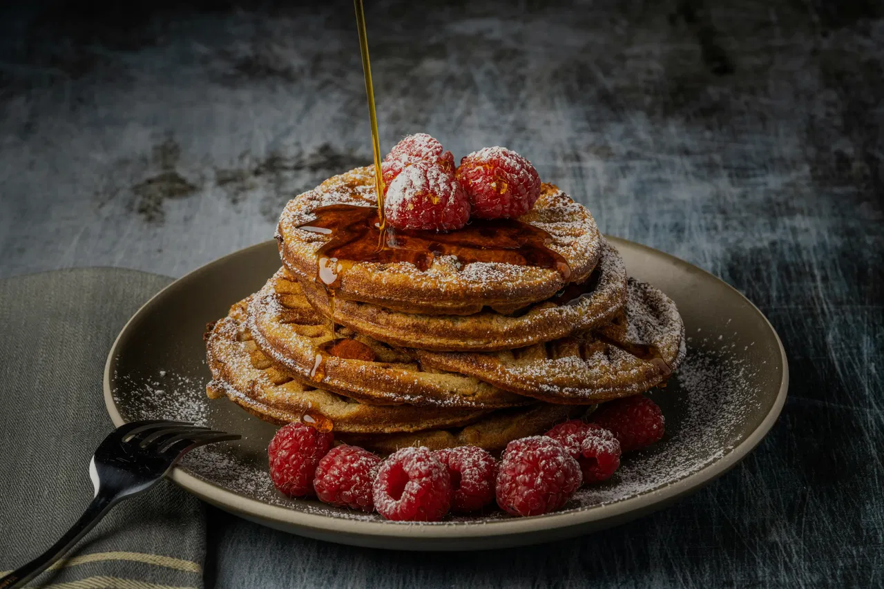 Stack of waffles with raspberries, syrup drizzle, and powdered sugar on a dark plate.