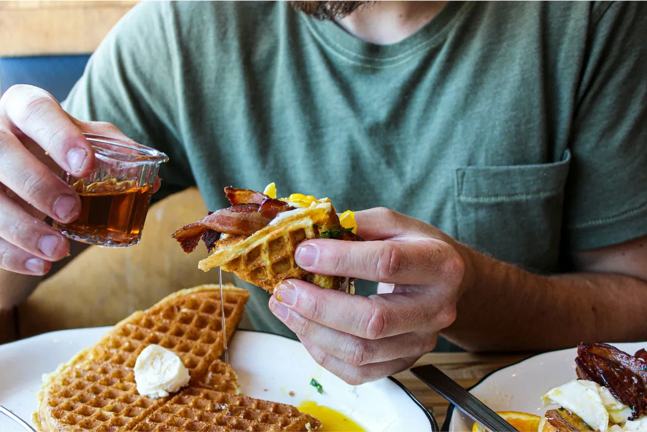 Man holding waffles with bacon, pouring syrup from a small cup over them.