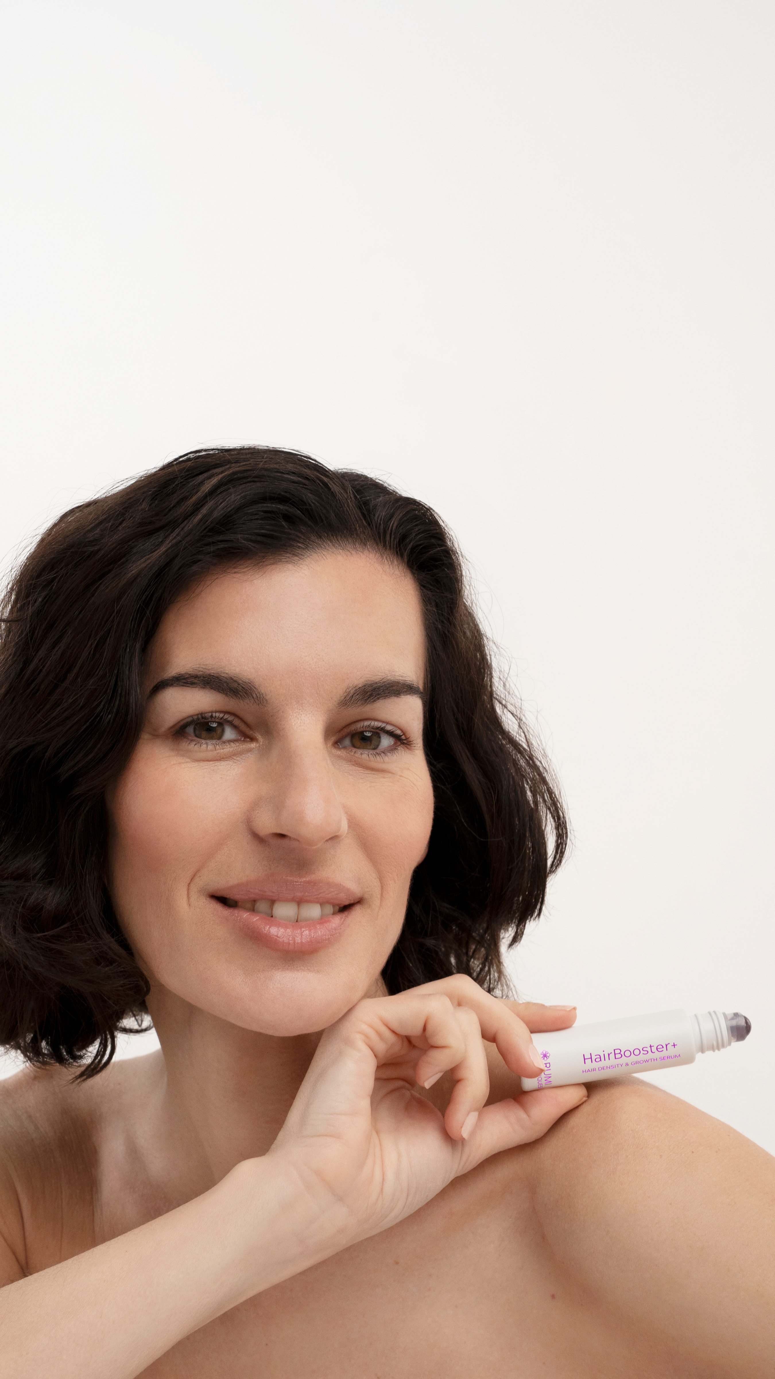 Smiling woman with a dark wavy bob holding the HairBooster+ roll-on serum by her chin