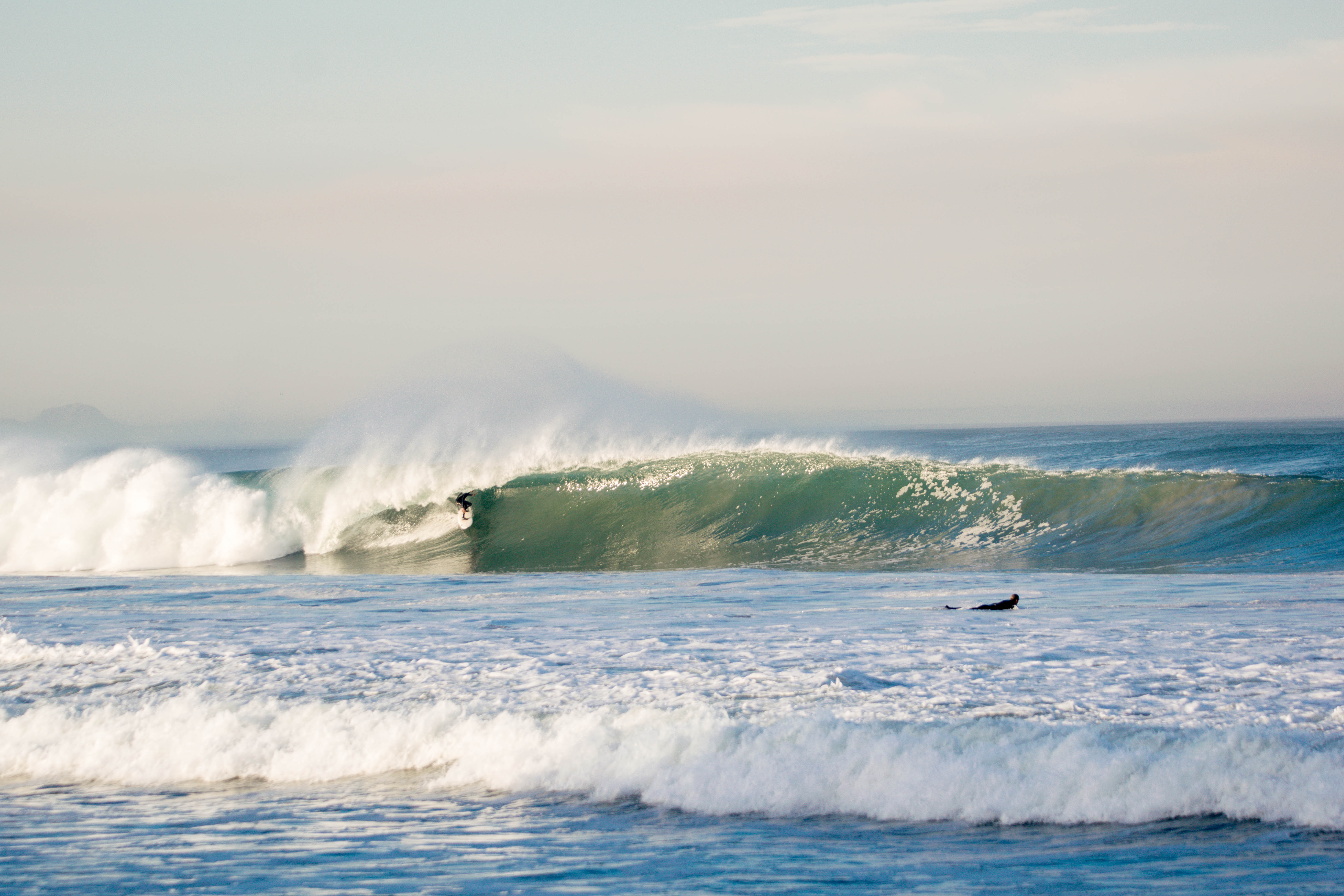 A surfer riding inside a breaking green wave while another paddles out