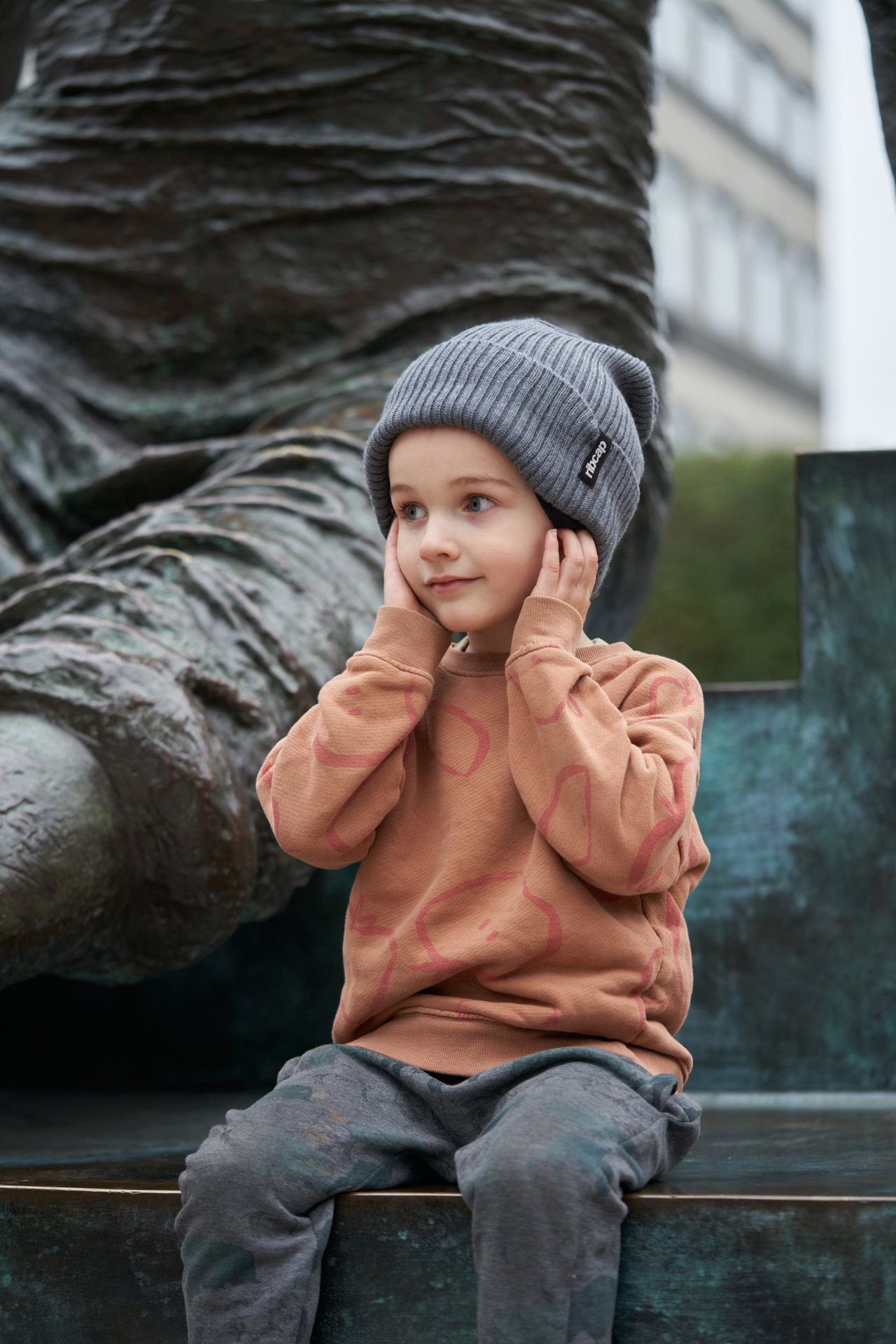 Young child wearing a gray beanie and brown sweater sitting on a bench with sculpture in the background