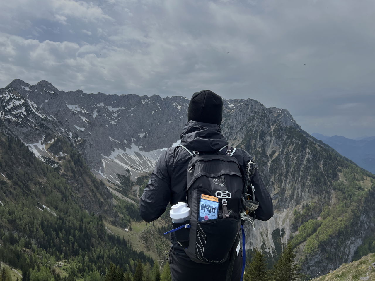 Person in black jacket and hat with backpack looks at rugged mountain landscape under cloudy sky featuring an insect protein sachet.