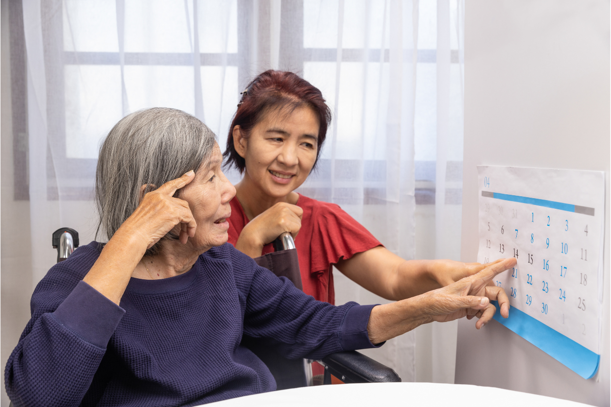 Person with dementia sitting in a wheelchair next to adult daughter looking at calendar with reminders.