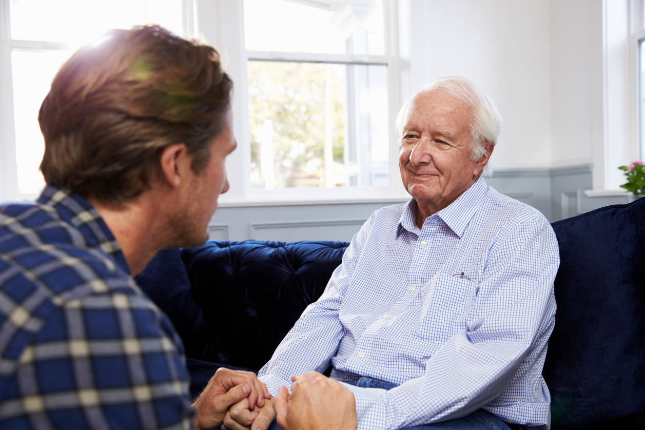Adult son holding hands with dad talking about Alzheimer's on a couch.