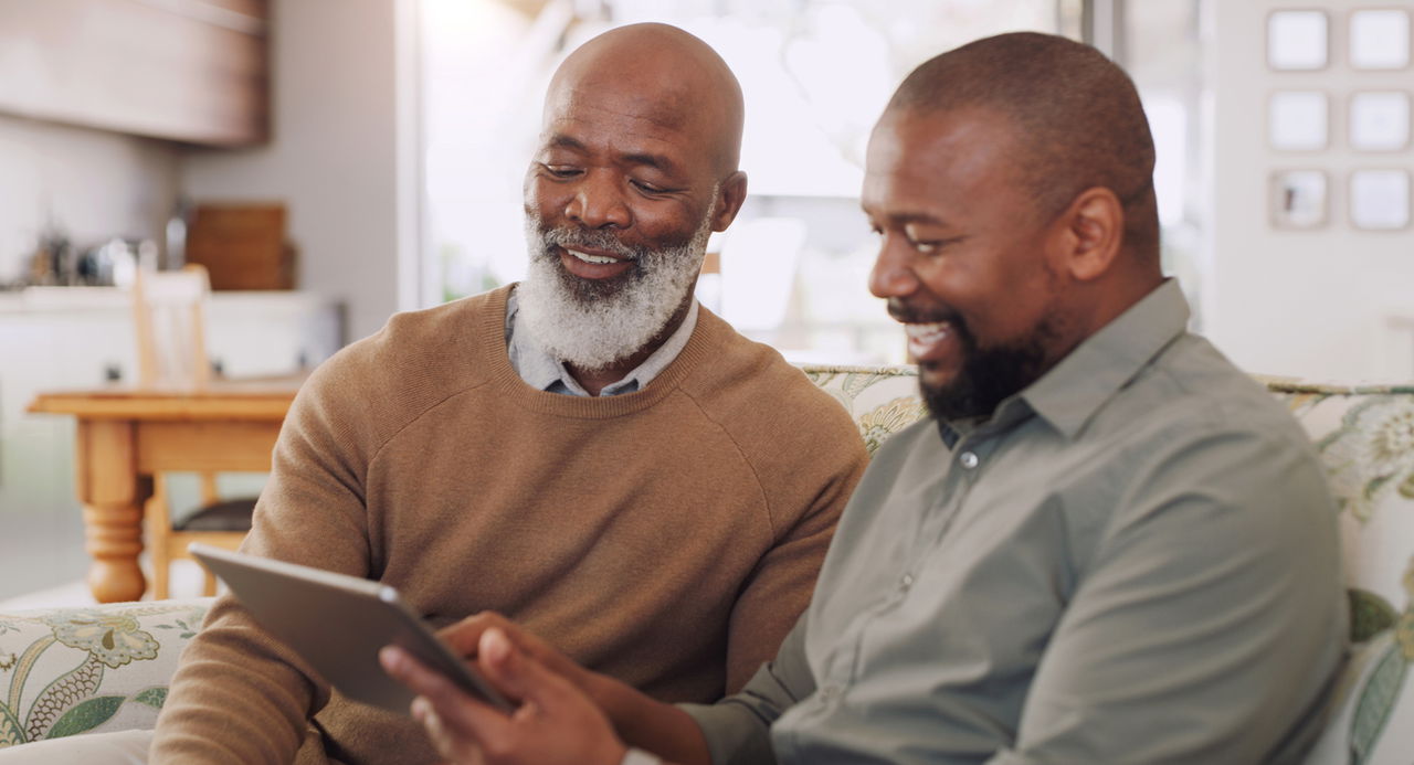 Father and adult son sitting on a couch looking at a message board tablet.