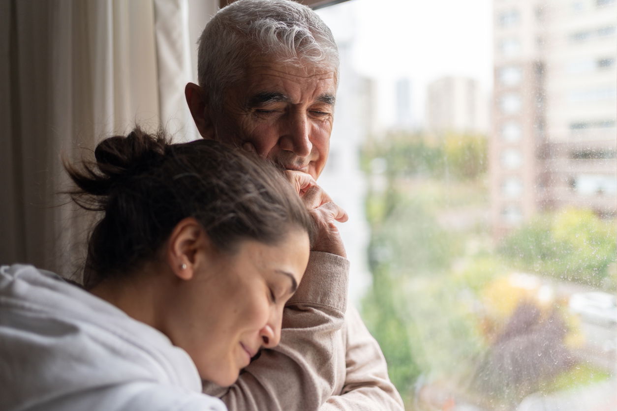 Older man and woman embrace peacefully by a window with city buildings in the background.