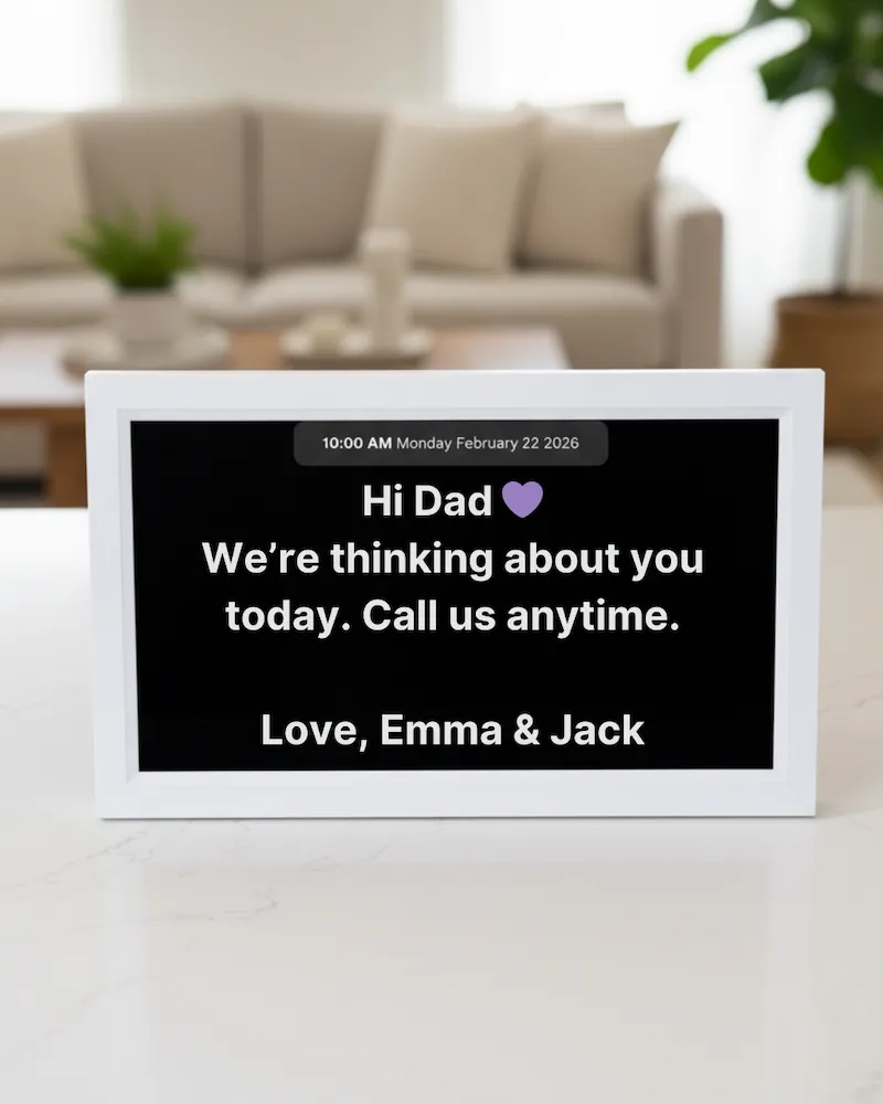 Memoryboard digital reminder display in a living room showing a message: "Hi Dad 💜 We're thinking about you today. Call us anytime. Love, Emma & Jack"