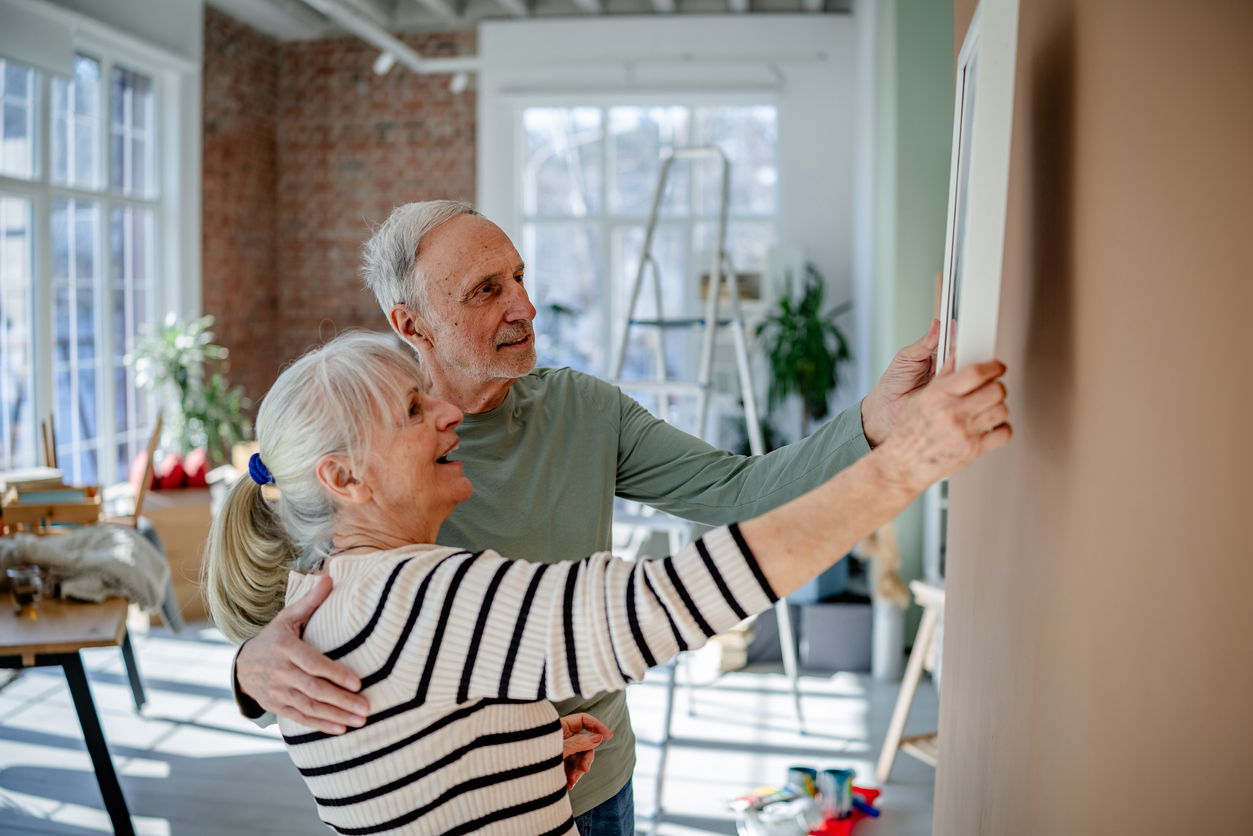 Elderly couple hanging a reminder on a wall in a sunlit room with a brick backdrop.