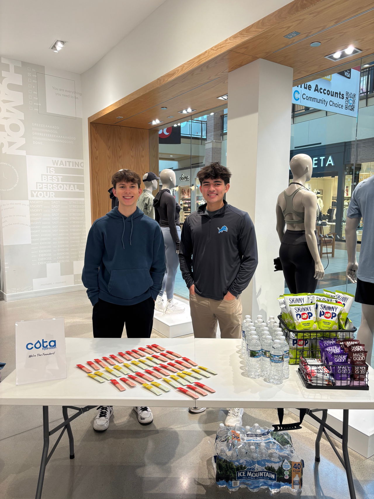 Two young men smiling behind a table with hydration sticks, water bottles, and snacks in a store.