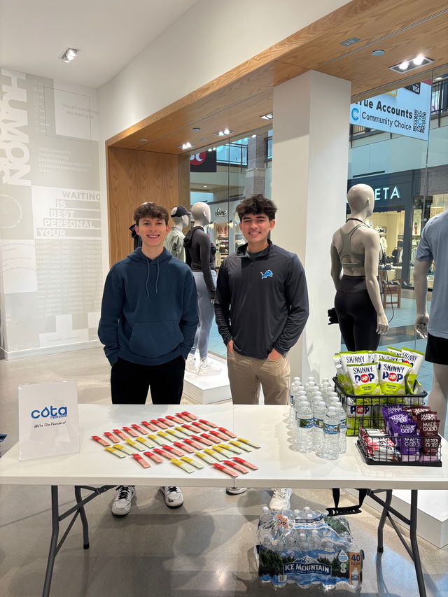 Two young men smiling behind a table with hydration sticks, water bottles, and snacks in a store.