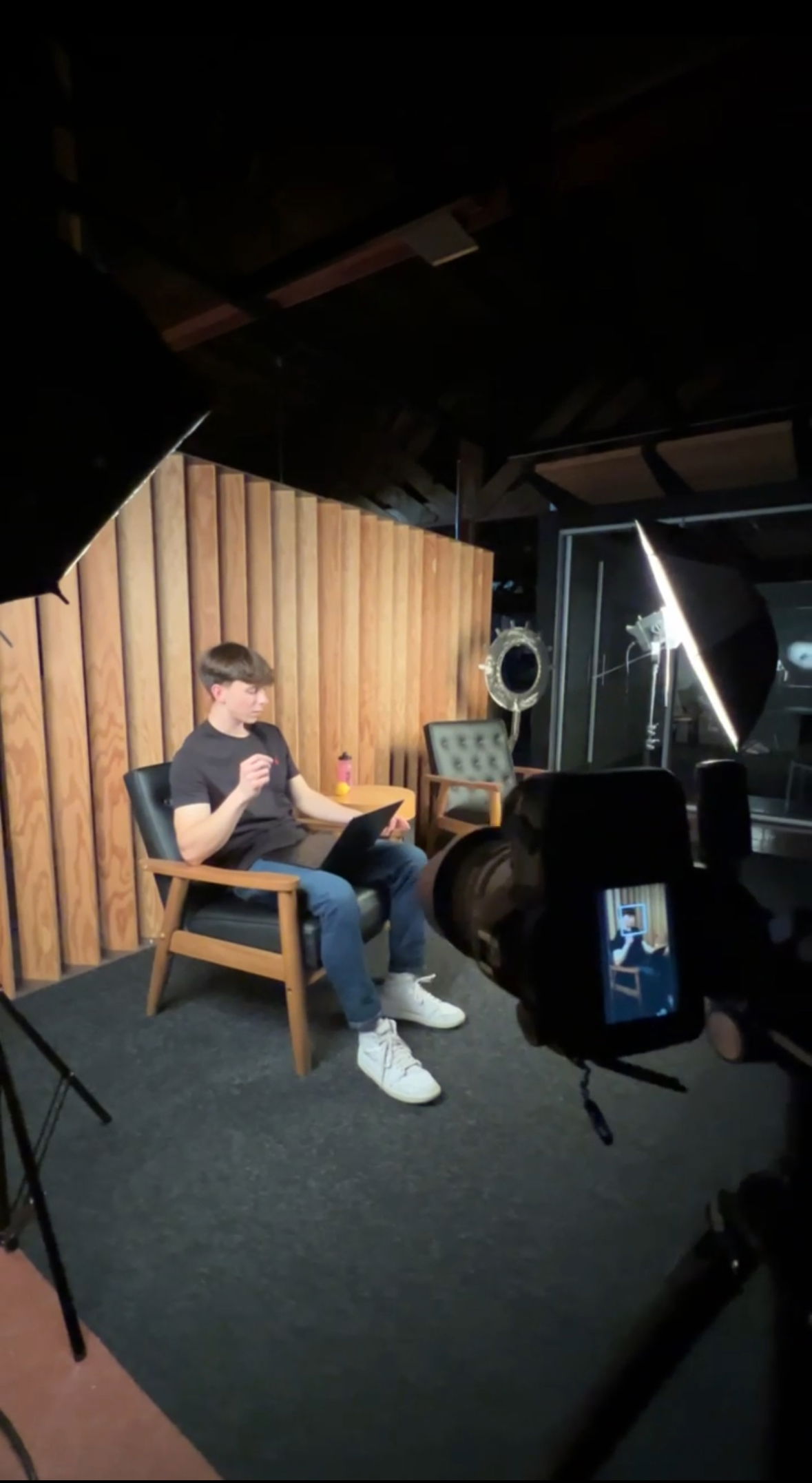 Young man sitting in chair with laptop, being recorded in a studio with professional lighting.