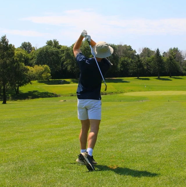 Golfer in navy shirt and white shorts swinging club on a sunny golf course.
