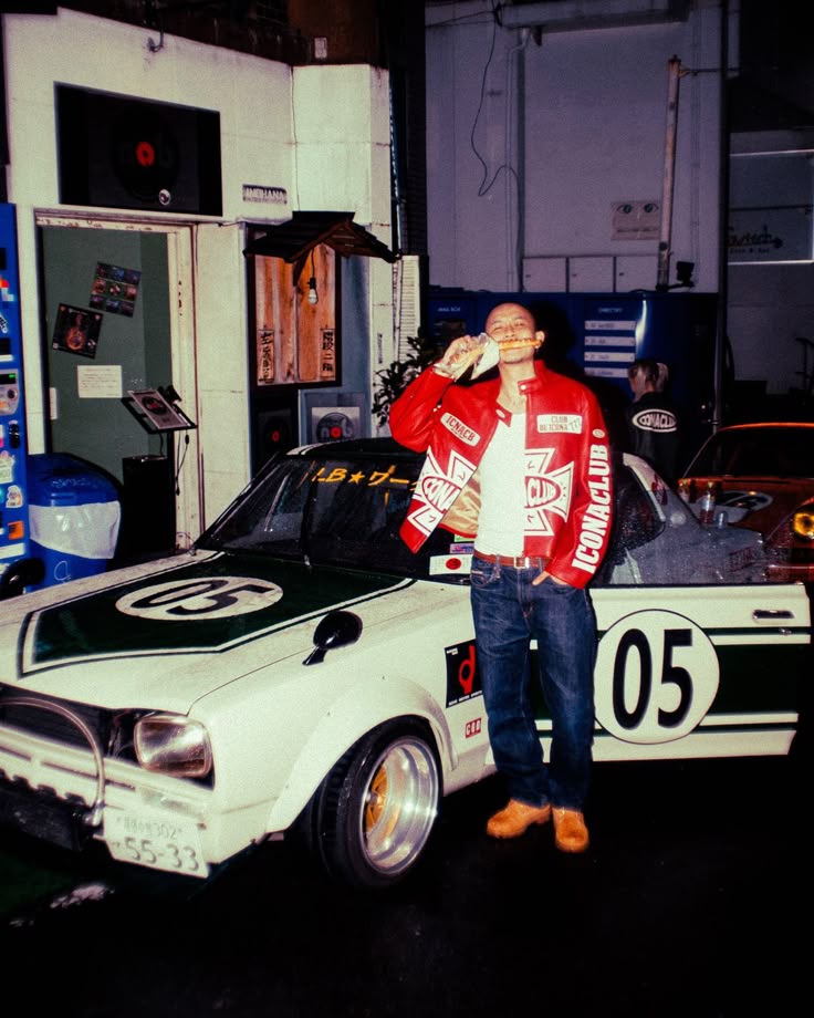 Racing jacket campaign image: a man in a red and white leather racing jacket, jeans and wheat boots standing in front of a vintage white race car numbered 05, shot with flash at night