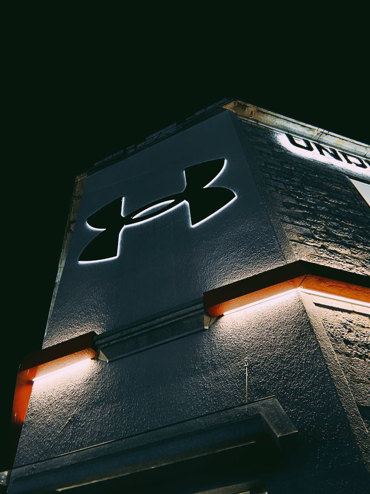 Under Armour campaign image: the backlit Under Armour logo glowing on the corner of a textured stone storefront at night, with orange light strips running along the ledges below