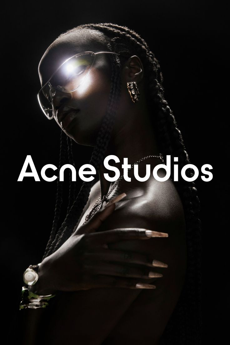 Acne Studios campaign portrait: a model with long braids wearing silver shield sunglasses, a fine chain necklace and stacked rings, lit by a single hard light against black