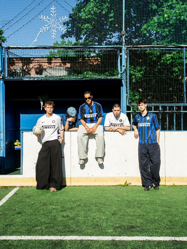 Sportswear campaign image: five friends in vintage Inter Milan Pirelli football shirts gathered by the dugout of a caged five-a-side pitch on a sunny day, one holding a football
