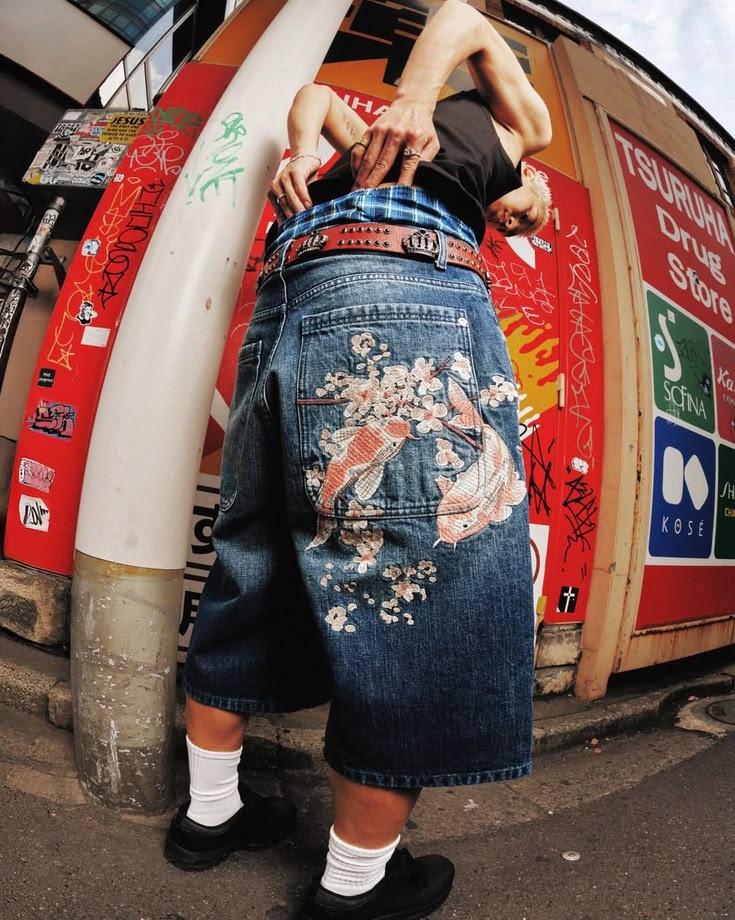 Derschutze campaign image: a fisheye shot from behind of baggy blue denim shorts with koi fish and cherry blossom embroidery across the back pocket, worn with a studded leather belt, white socks and black shoes in front of a graffiti-covered red storefront