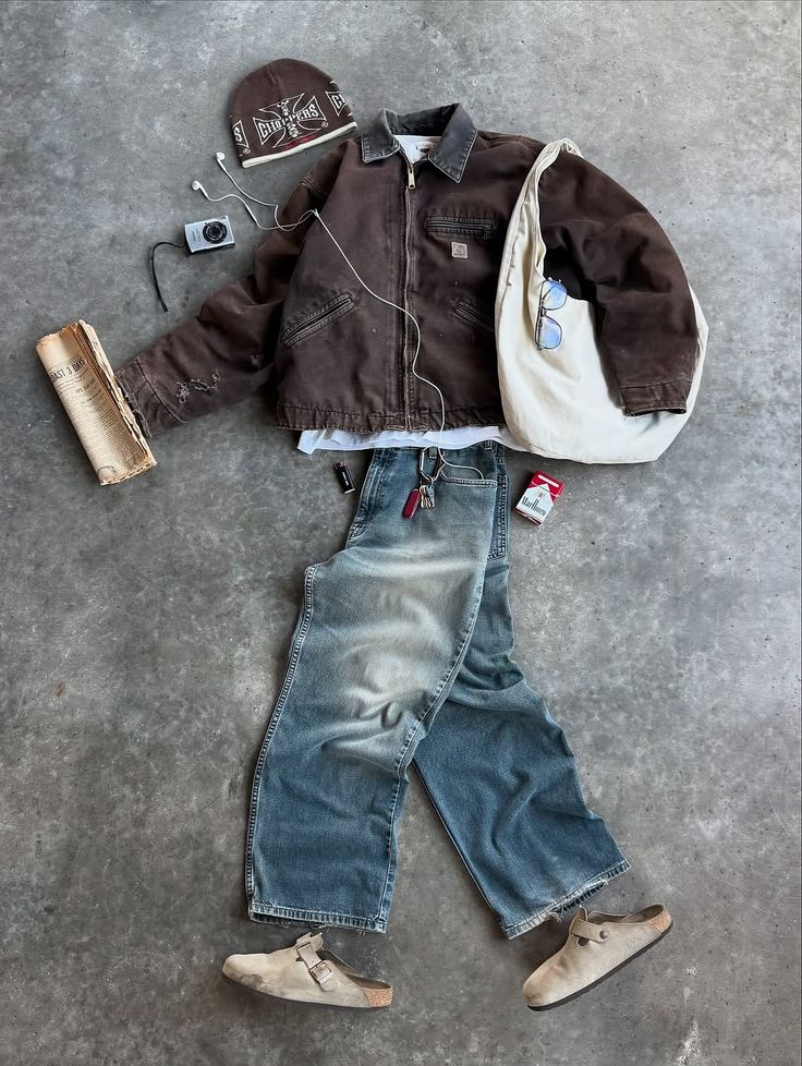 Flat-lay of a full vintage outfit on concrete: brown work jacket, faded baggy jeans, beanie, suede clogs and a canvas shoulder bag