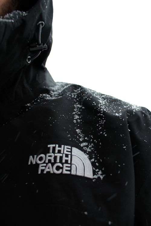 The North Face campaign image: a close-up of a black hooded North Face shell jacket dusted with falling snow, the white embroidered Half Dome logo on the chest