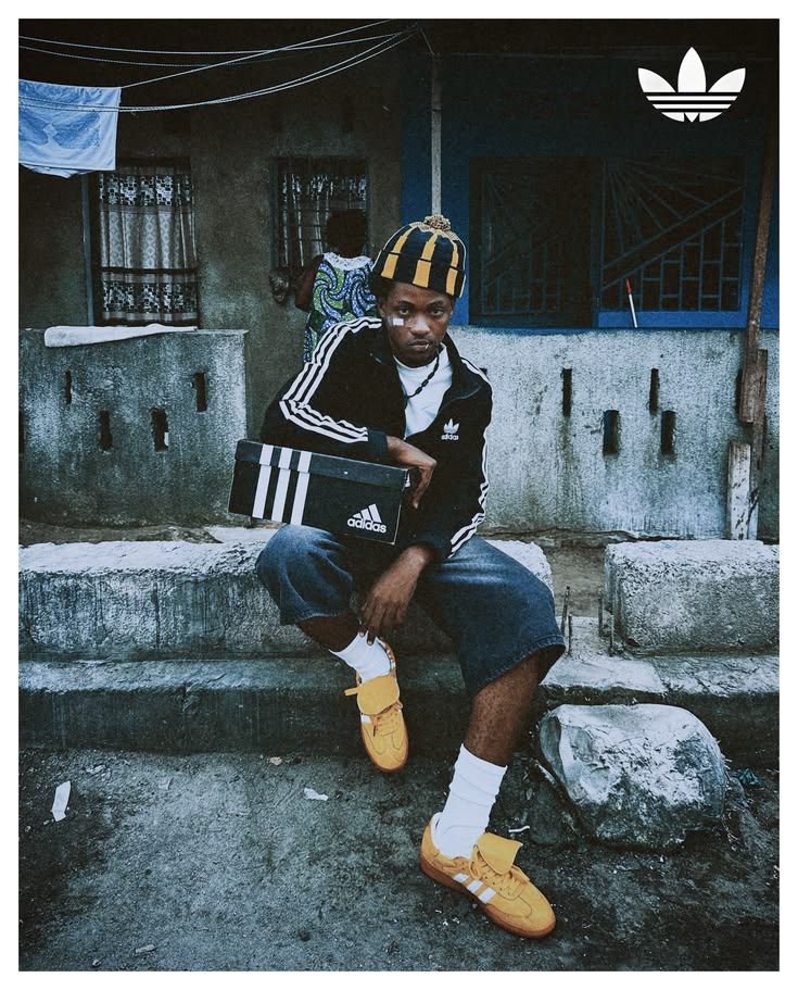 Adidas campaign portrait: a man in a black Adidas tracksuit jacket, striped beanie and yellow Sambas sitting on a concrete step with an Adidas shoe box on his lap