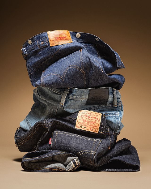 Levi's campaign image: a stack of folded vintage Levi's 501 jeans in raw, faded and rinse indigo denim, showing the Two Horse leather patches and a Red Tab against a warm tan studio backdrop