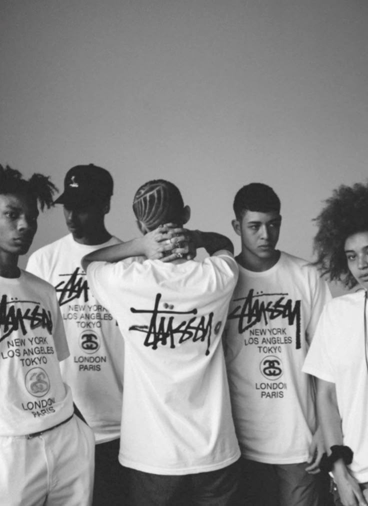 Stüssy campaign image: a figure in a navy workwear set standing in shallow water under a concrete bridge, their shadow on the wall holding a saxophone