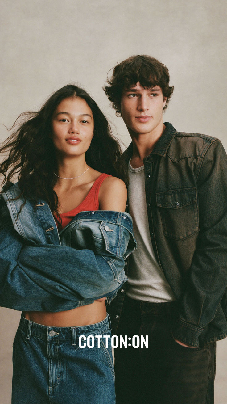 Cotton On campaign portrait: a woman in a red tank top with a light-wash denim jacket slipped off her shoulders and a man in a washed black denim western shirt over a grey tee, posed together against a warm grey studio backdrop