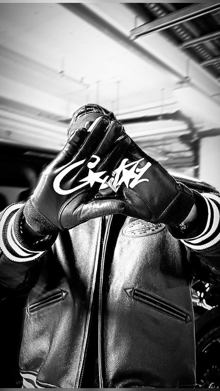 Corteiz campaign image: a figure in a black leather varsity jacket with striped cuffs raising both hands in black leather gloves printed with the white CRTZ logo, shot in black and white